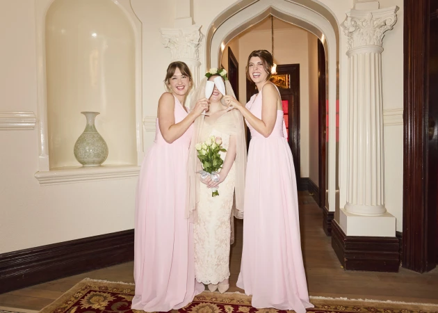Bride with bridesmaids in pink dresses holding flowers in indoor venue with arched doorway and columns, celebrating wedding day.
