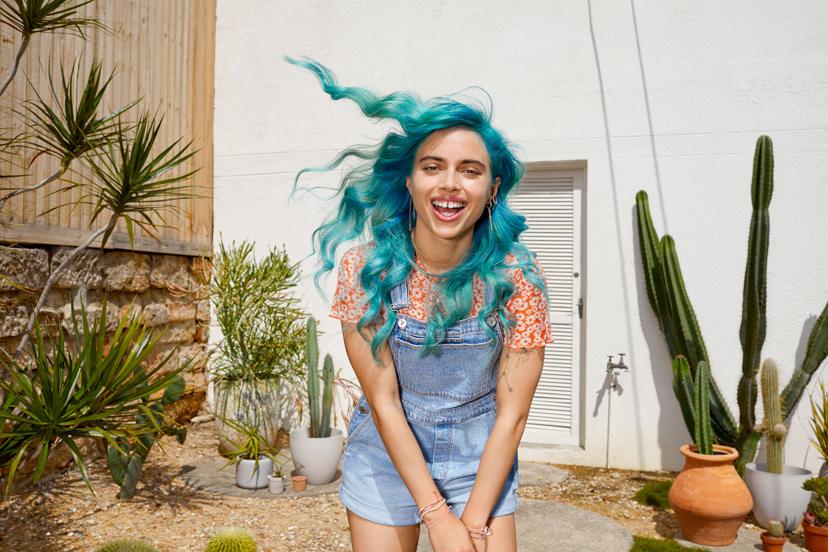 Smiling woman with blue hair poses in denim overalls, surrounded by potted cacti and desert plants in a bright outdoor setting.