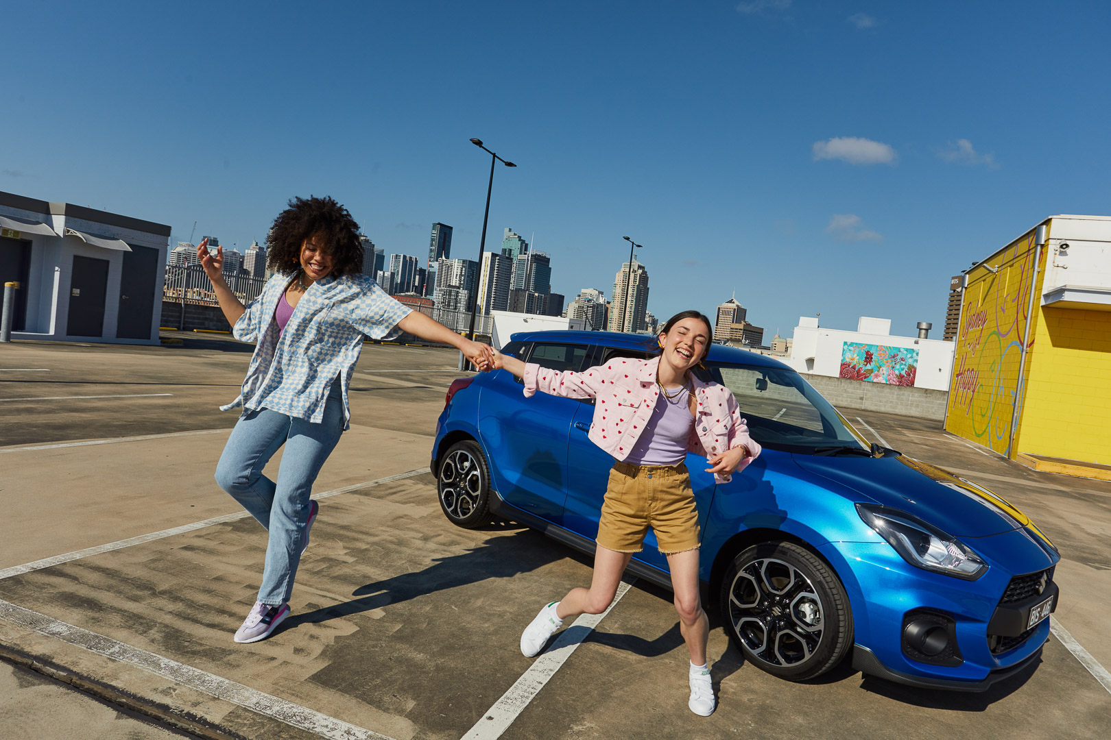 Two young women joyfully pose beside a blue Suzuki Swift car on a rooftop parking lot, city skyline in the background, bright sunny day.