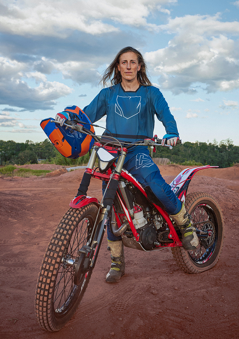 Female athlete poses confidently with a red-framed trial motorcycle on a dirt track under a cloudy sky in an eye-level medium shot.