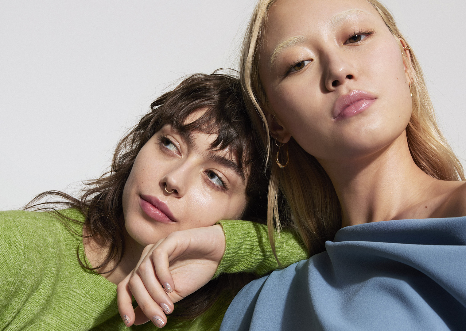 Two women lean against each other indoors. One woman wears a green sweater and the other a blue top against a white background.