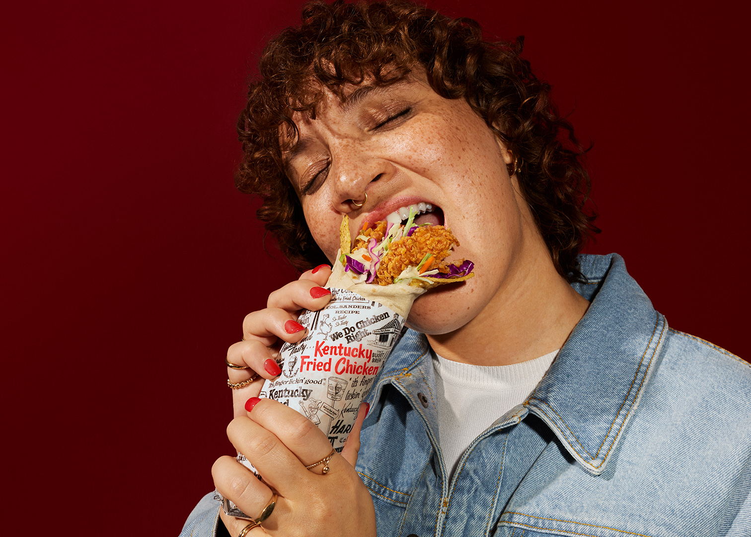Woman with curly hair eating a chicken wrap on a red background. Hands holding the wrap are adorned with gold rings.