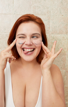 Close-up of a smiling woman with red hair and under-eye masks, wearing a white top, posing indoors against a tiled wall.