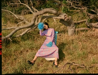 Woman in pink floral dress with blue feather cuffs seated outdoors on a chair beneath large fallen tree branches in grassy terrain.