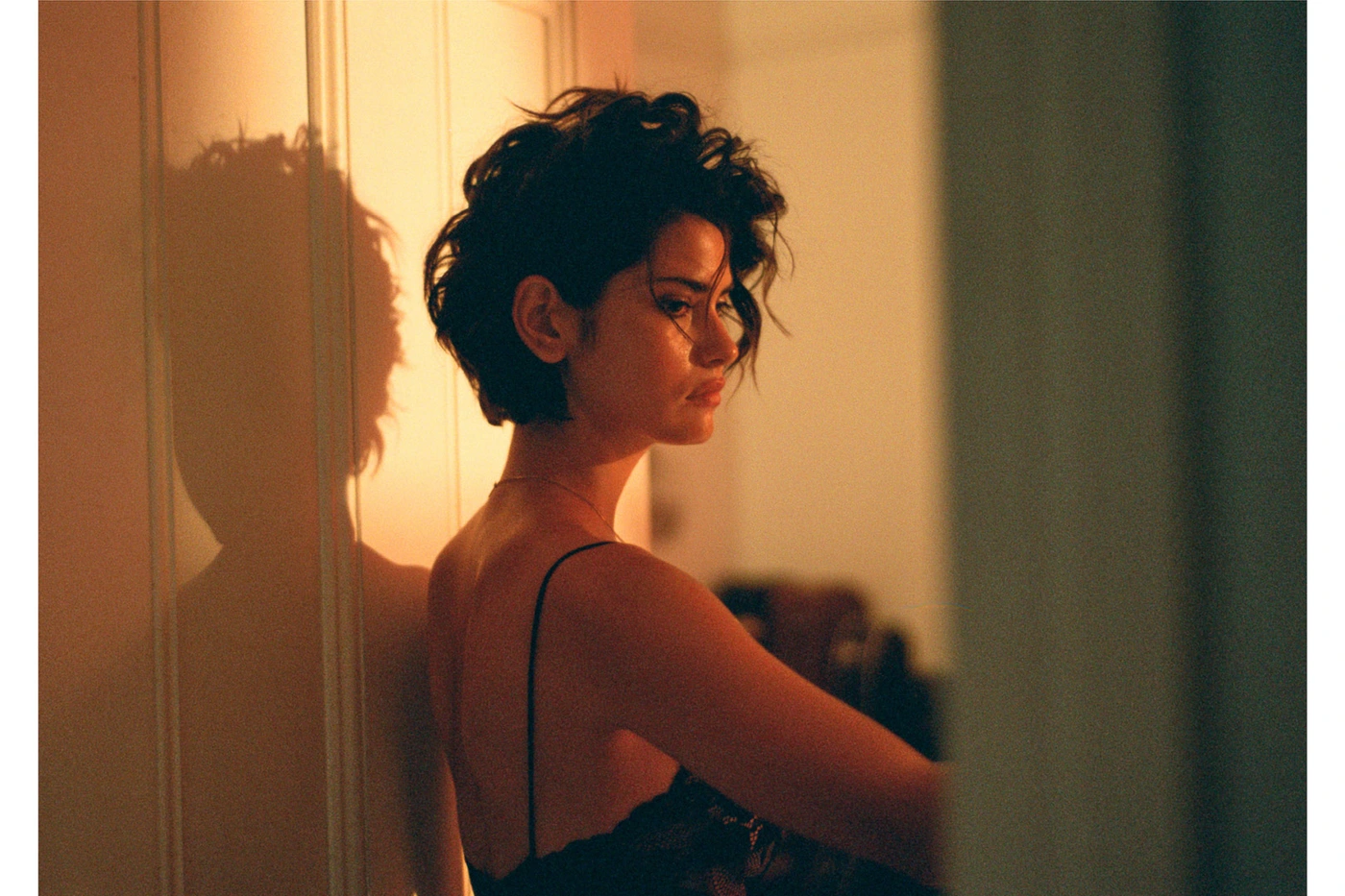 A woman with short dark curly hair wearing a dark strap top, casting a silhouette shadow on a warm lit wall by Sarah Adamson.