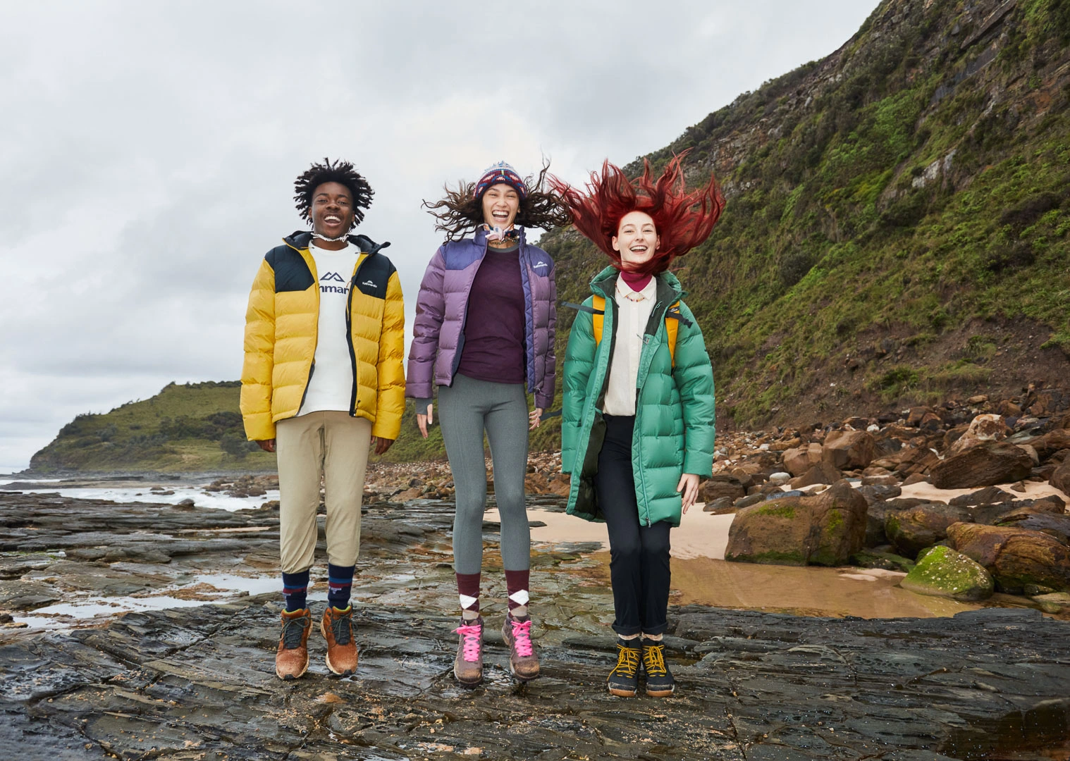 Three young friends jumping together on a rocky coastal shore wearing colorful outdoor winter jackets and hiking boots.