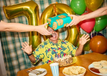 Elderly man in yellow floral shirt with a birthday cake and foil number balloons by Sarah Adamson