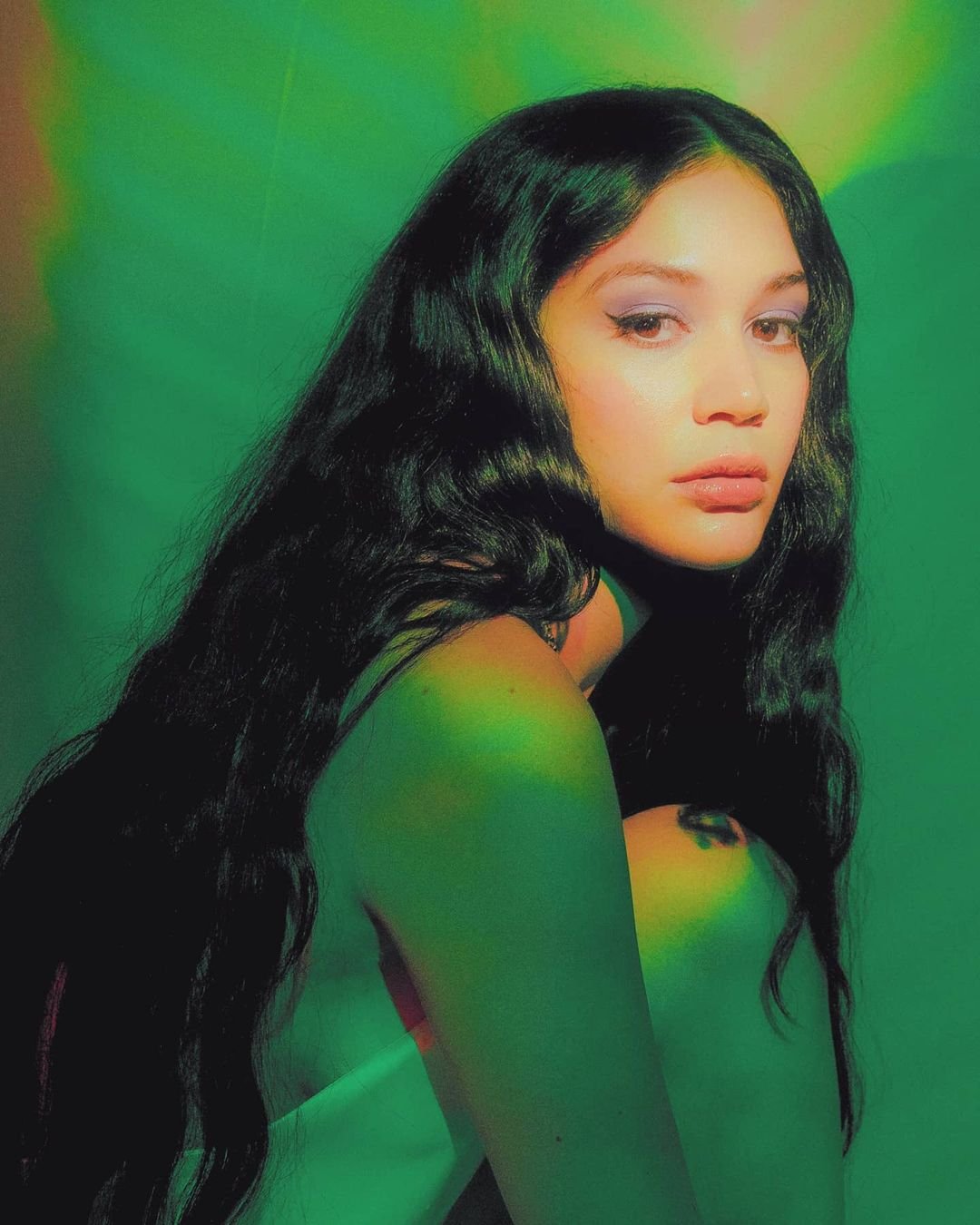 Close-up studio portrait of a woman with long dark hair and light skin, wearing makeup, against a green background with soft lighting.
