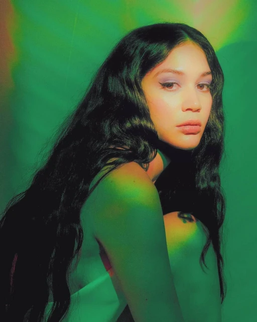 Close-up studio portrait of a woman with long dark hair and light skin, wearing makeup, against a green background with soft lighting.