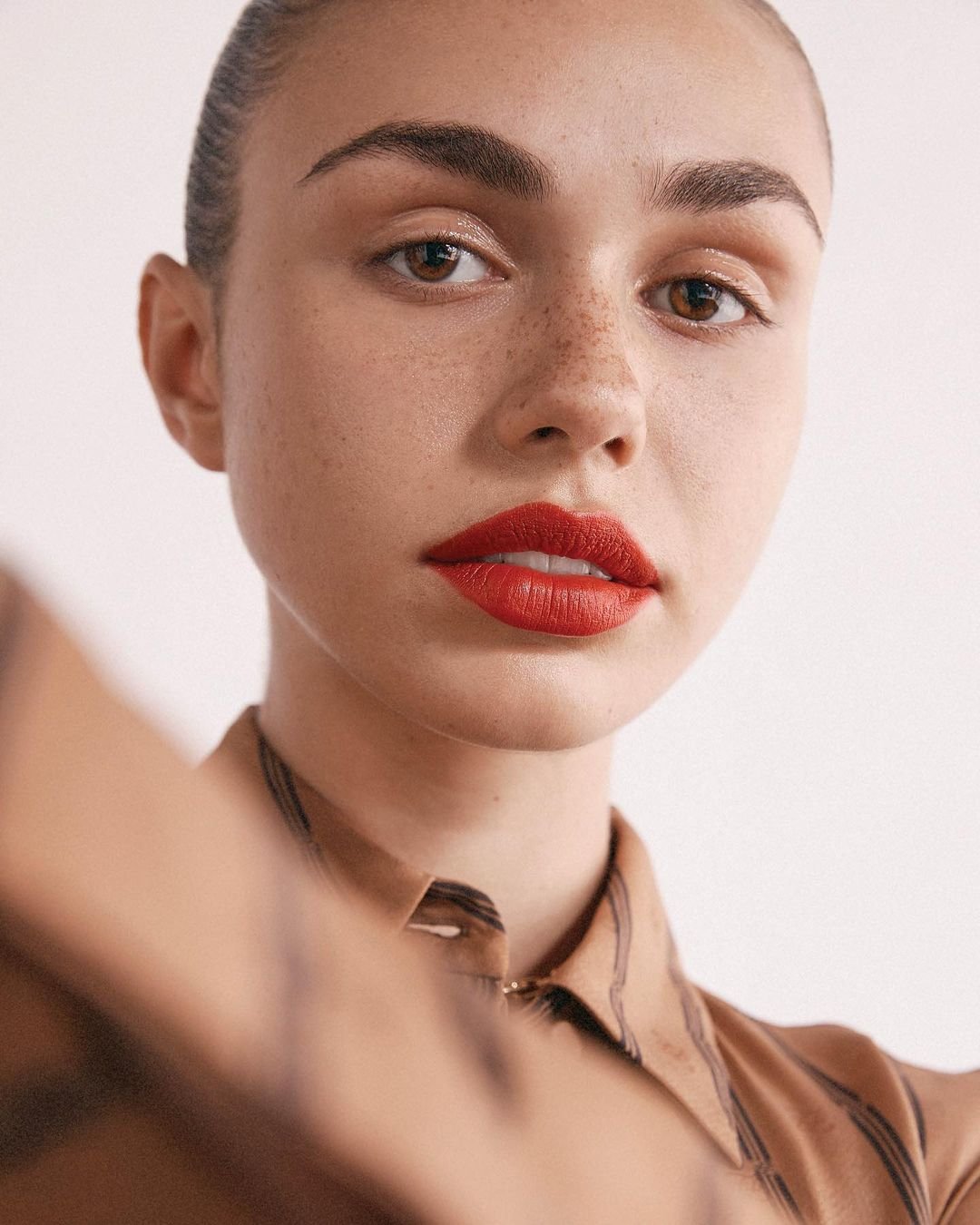 Close-up portrait of a light-skinned woman with red lipstick and freckles wearing a patterned blouse against a plain white background.