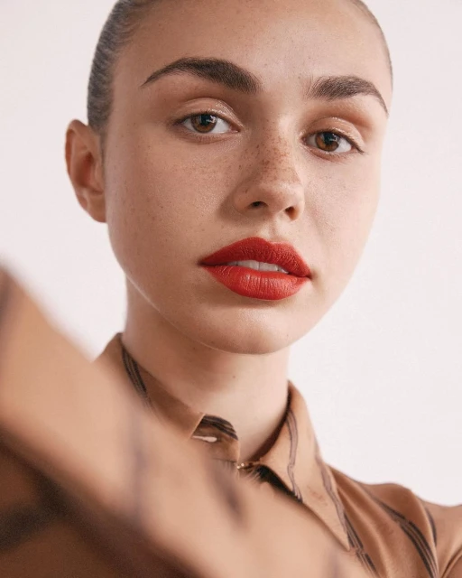 Close-up portrait of a light-skinned woman with red lipstick and freckles wearing a patterned blouse against a plain white background.