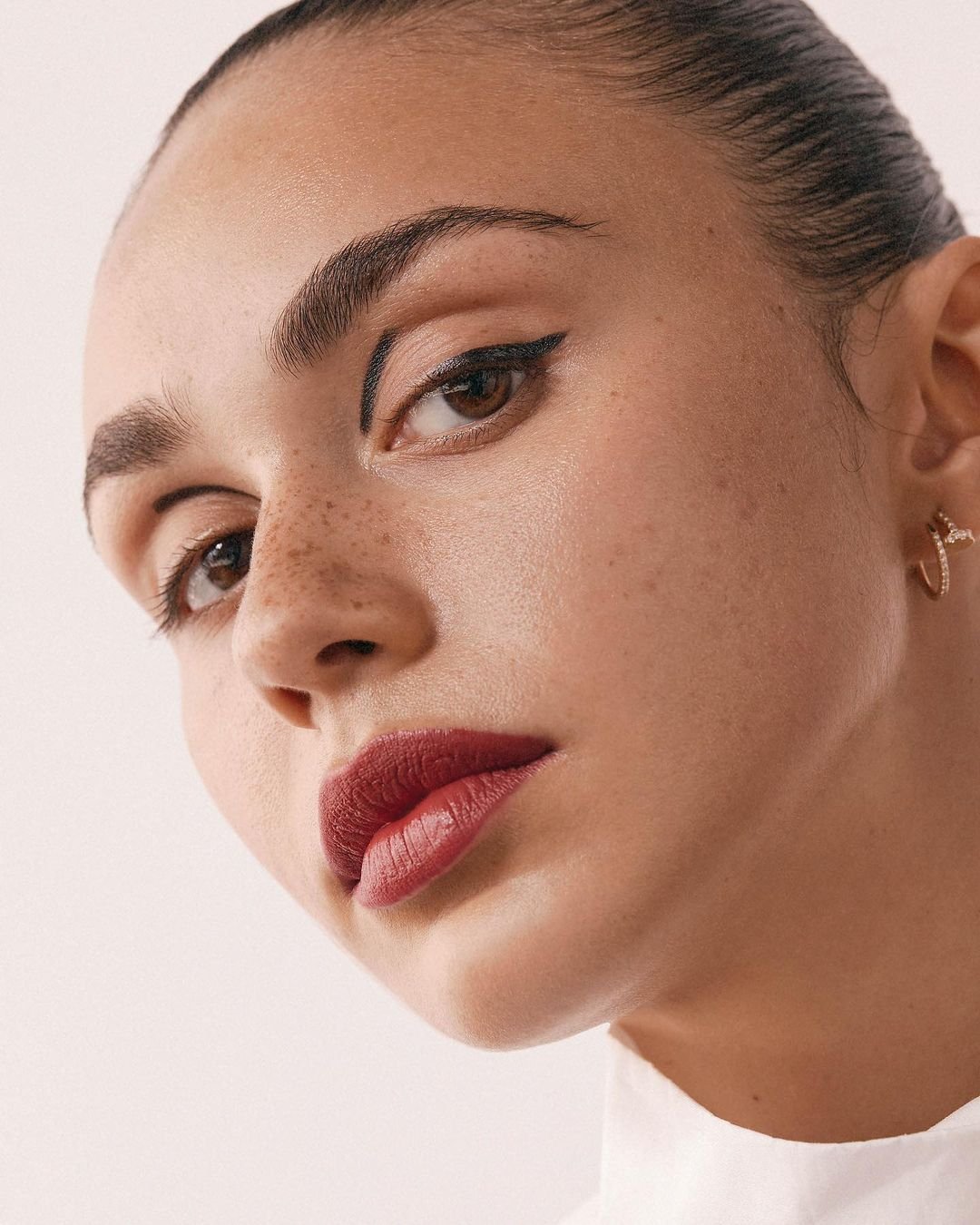 Close-up of a young woman with brown eyes, wearing red lipstick and gold hoop earrings, against a light-colored backdrop. Beauty shot.