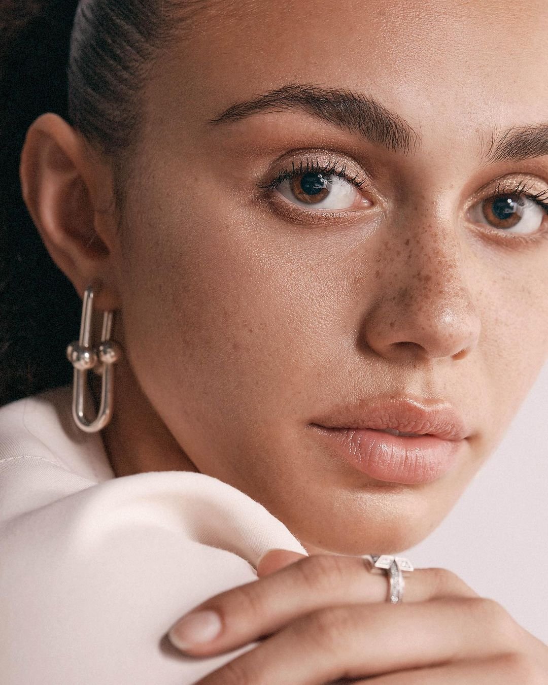 Close-up portrait of a woman with silver earrings and a diamond ring, focusing on her face and jewelry against a light background.