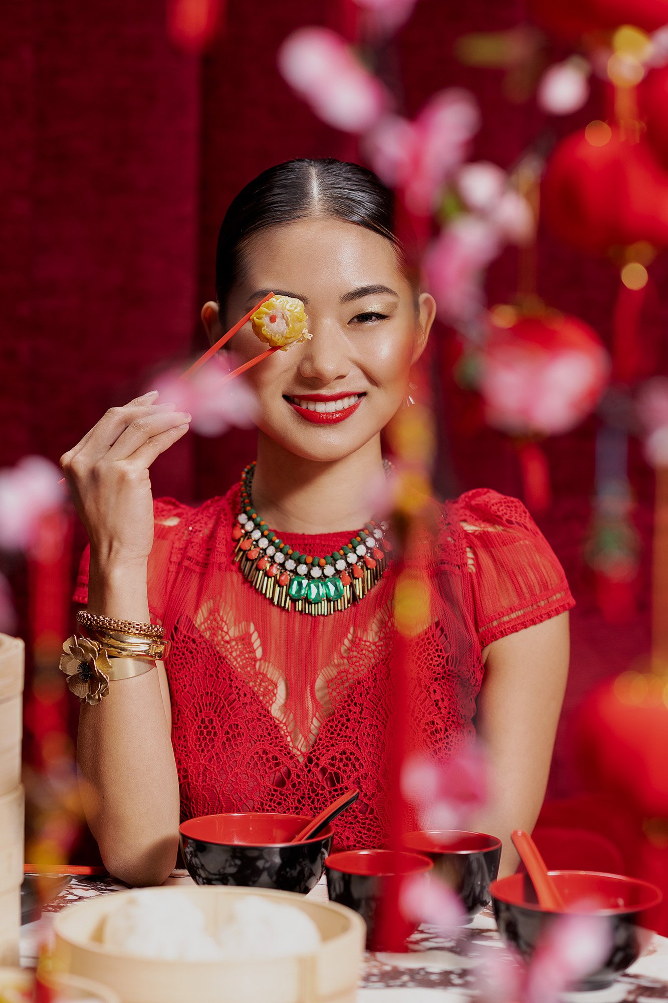 Smiling Asian woman in red dress holds dim sum with chopsticks, celebrating Chinese new year with traditional decorations and food.