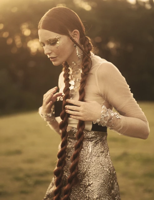Woman in a shimmering gold dress with long auburn braids and glitter makeup poses in a grassy outdoor setting during golden hour.