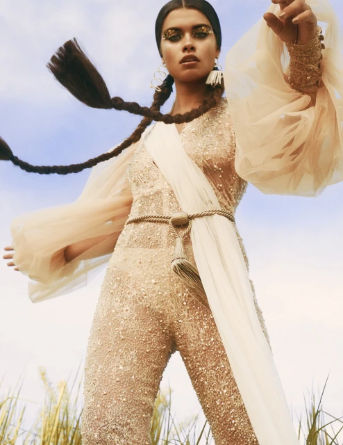 Brunette woman poses outdoors in a gold jumpsuit and long braids against a blue sky and field.