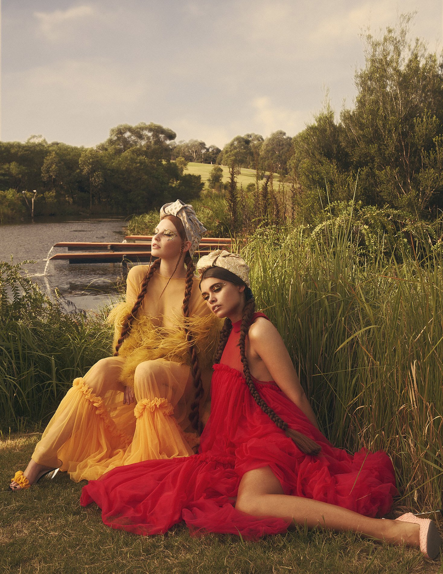 Two models in sheer, colorful dresses pose in a grassy field near a pond, creating a whimsical and fashionable outdoor scene.