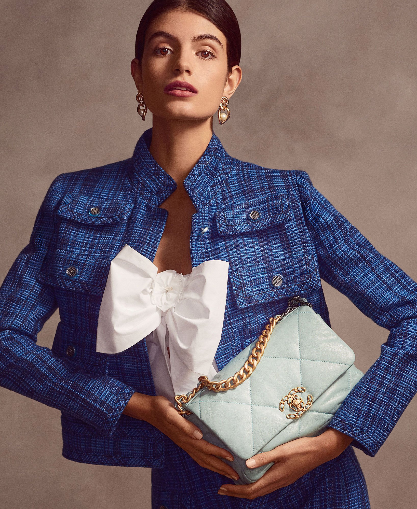 Brunette woman poses with a light blue handbag and blue tweed jacket, adorned with a white bow blouse and gold jewelry. Fashion shot.