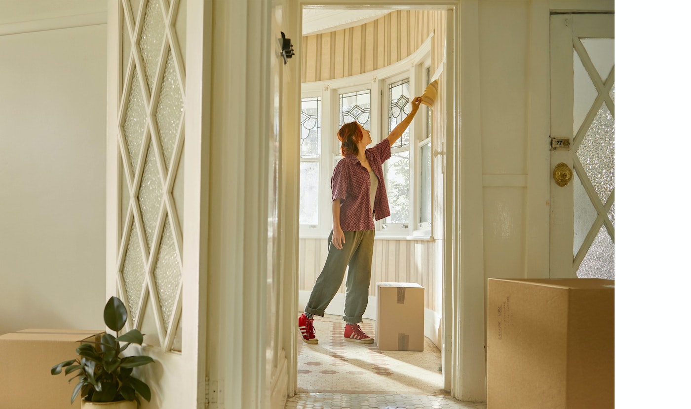 Woman in a casual outfit renovates a home interior by painting a room with white walls, tile floor, and boxes in the hallway.