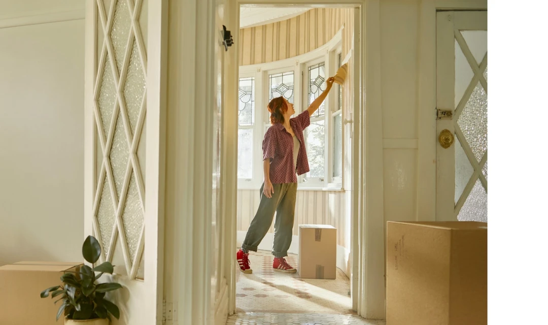 Woman in a casual outfit renovates a home interior by painting a room with white walls, tile floor, and boxes in the hallway.
