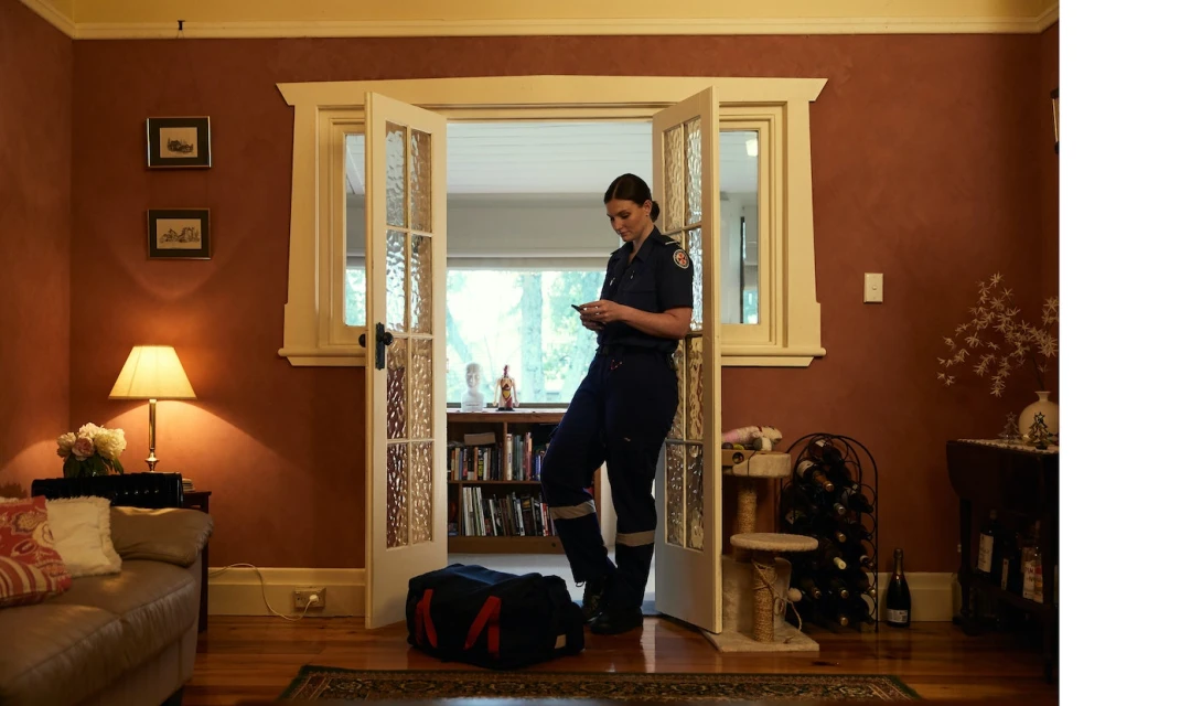 Female paramedic in uniform stands in doorway, looking at her smartphone with medical bag nearby in a home interior setting.