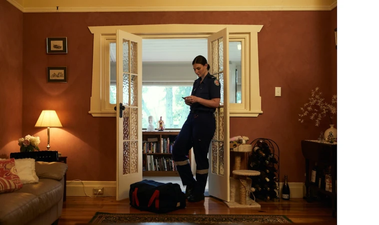 Female paramedic in uniform stands in doorway, looking at her smartphone with medical bag nearby in a home interior setting.