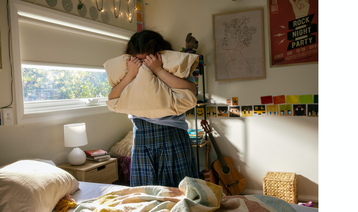 A person holding a pillow in a sunlit bedroom, featuring a bed, window, wall decorations, and guitar, capturing an intimate indoor scene.
