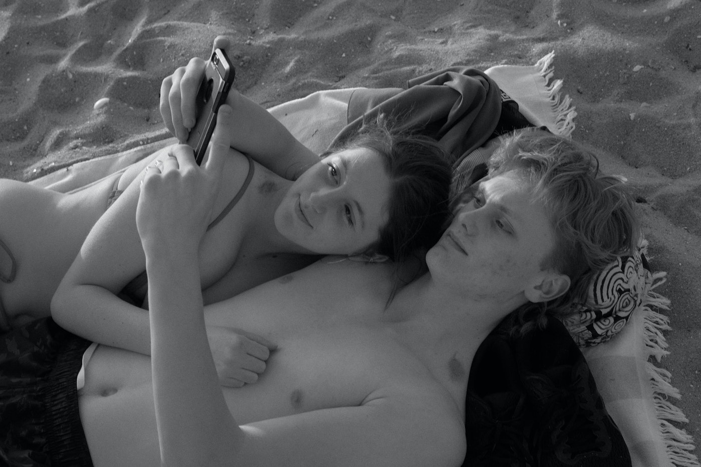 Black and white photo of a young couple lying on a blanket on a sandy beach, one holding a smartphone, capturing a relaxed, intimate moment.