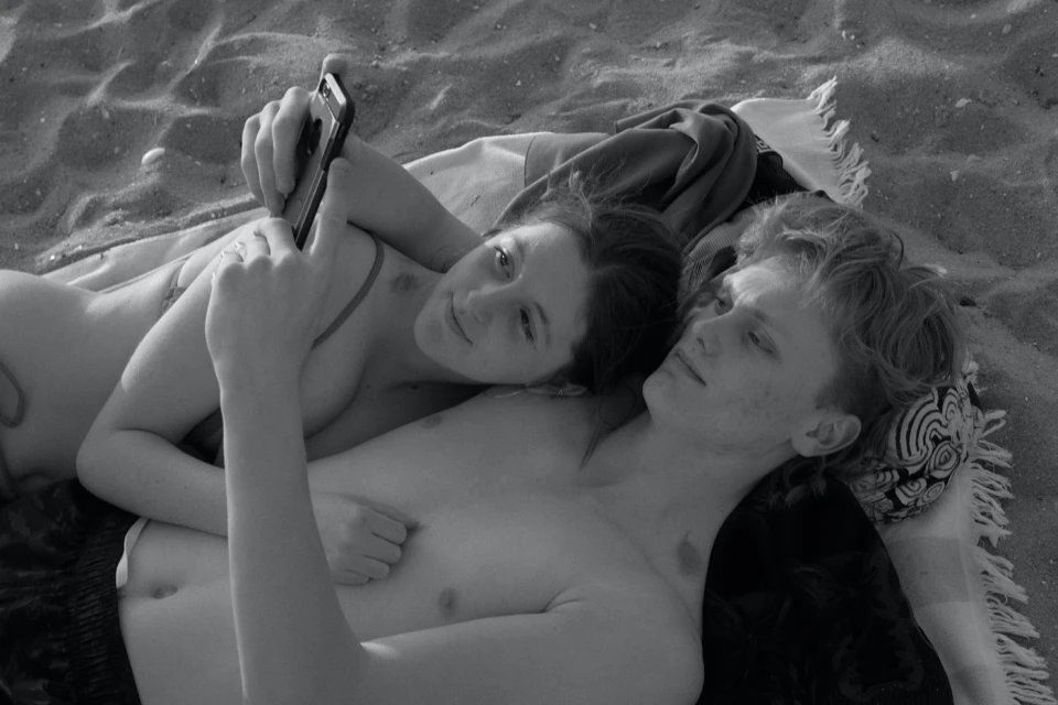 Black and white photo of a young couple lying on a blanket on a sandy beach, one holding a smartphone, capturing a relaxed, intimate moment.
