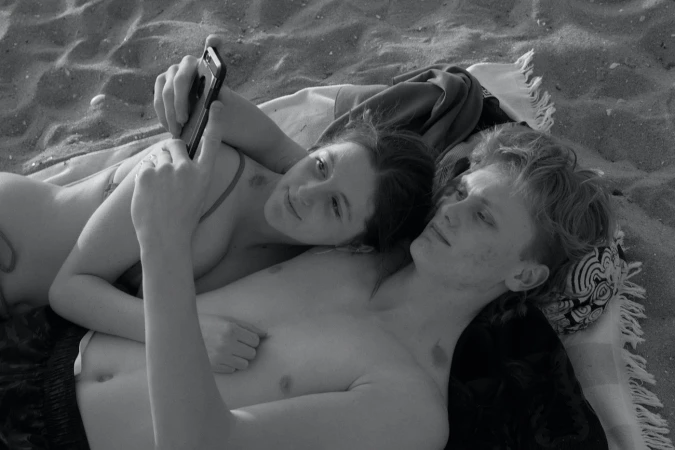 Black and white photo of a young couple lying on a blanket on a sandy beach, one holding a smartphone, capturing a relaxed, intimate moment.