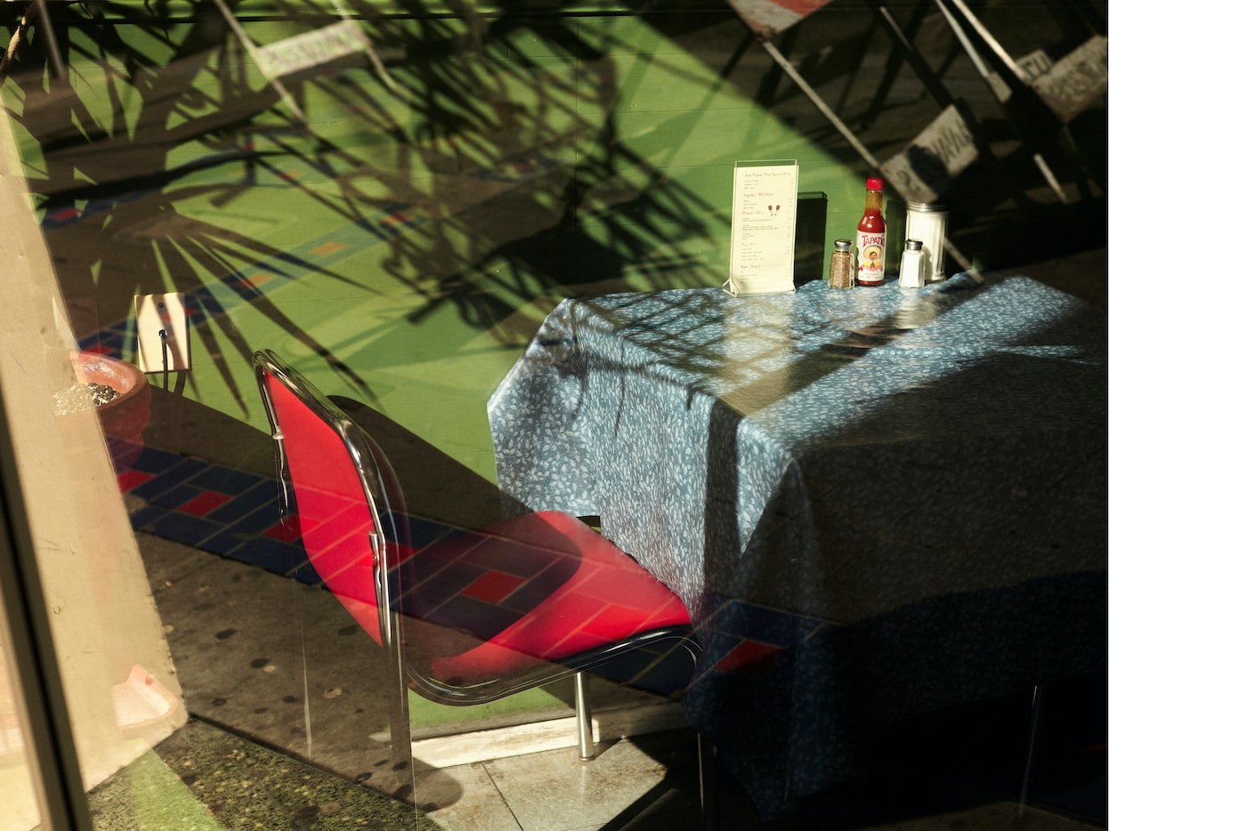 Empty red chair at a table with a blue tablecloth in a restaurant with window reflections and interior shadows creating a quiet scene.