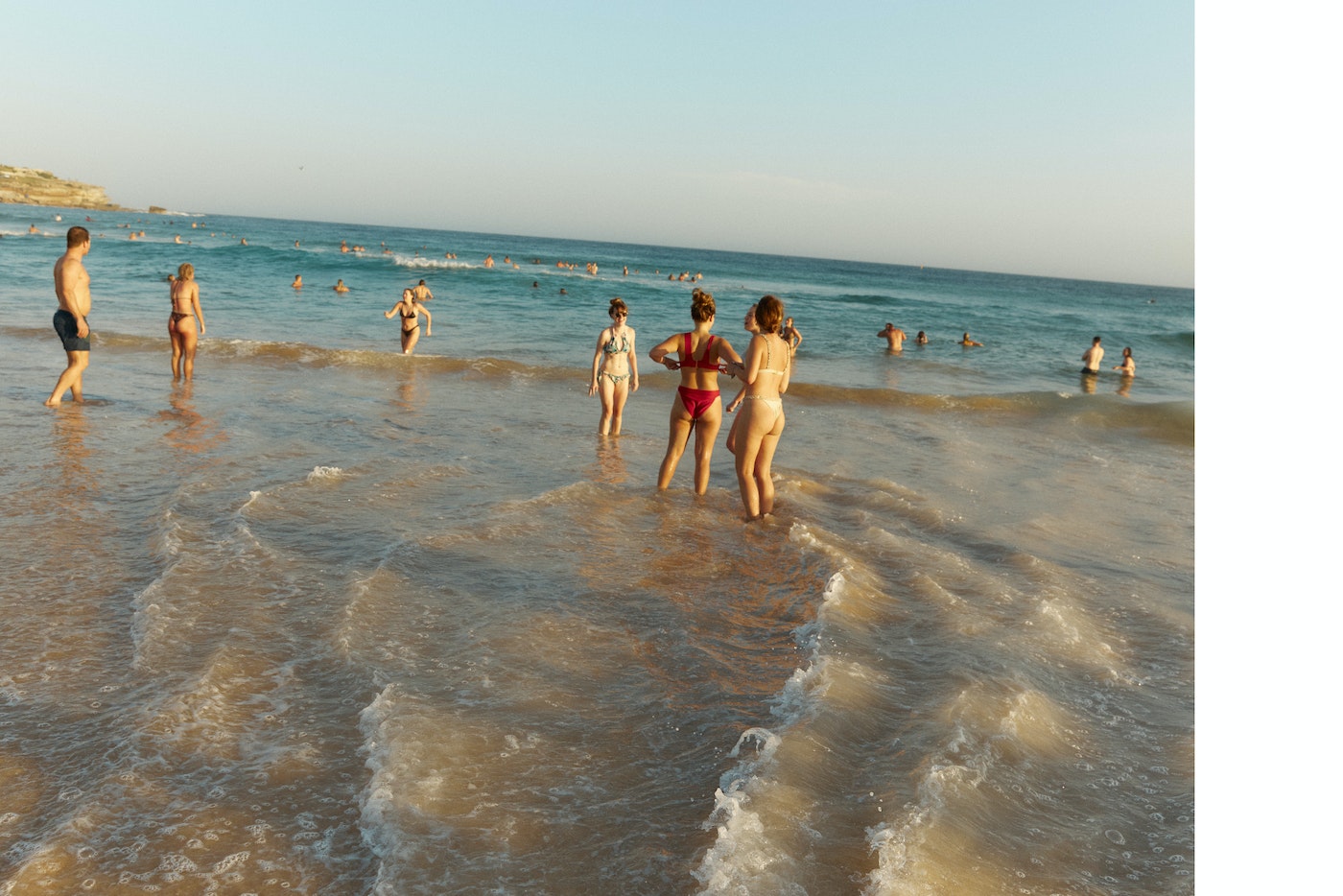 People swimming and wading in shallow ocean surf along a sandy beach under a clear sky.
