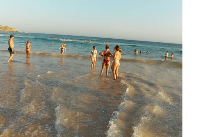 People swimming and wading in shallow ocean surf along a sandy beach under a clear sky.