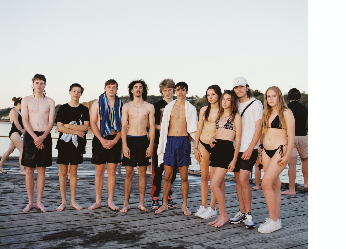 Group of young people in swimwear stand on a wooden dock by a lake. Some are shirtless and wearing shorts; it's a sunny, casual scene.