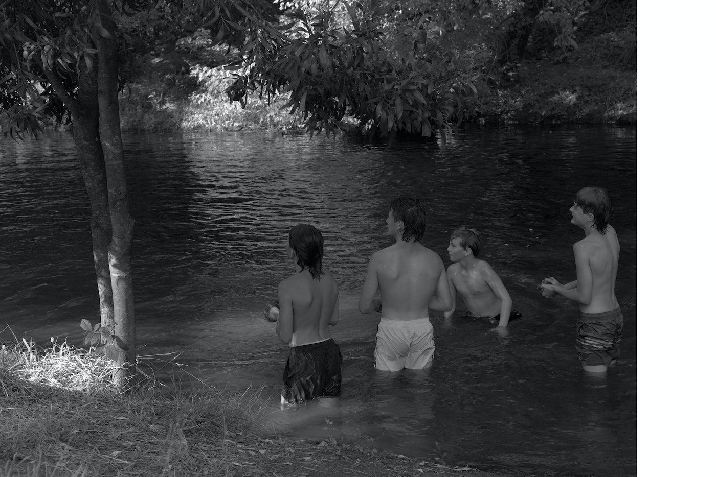 Four youths stand in a river surrounded by trees and vegetation, captured in a monochromatic image with a documentary feel.