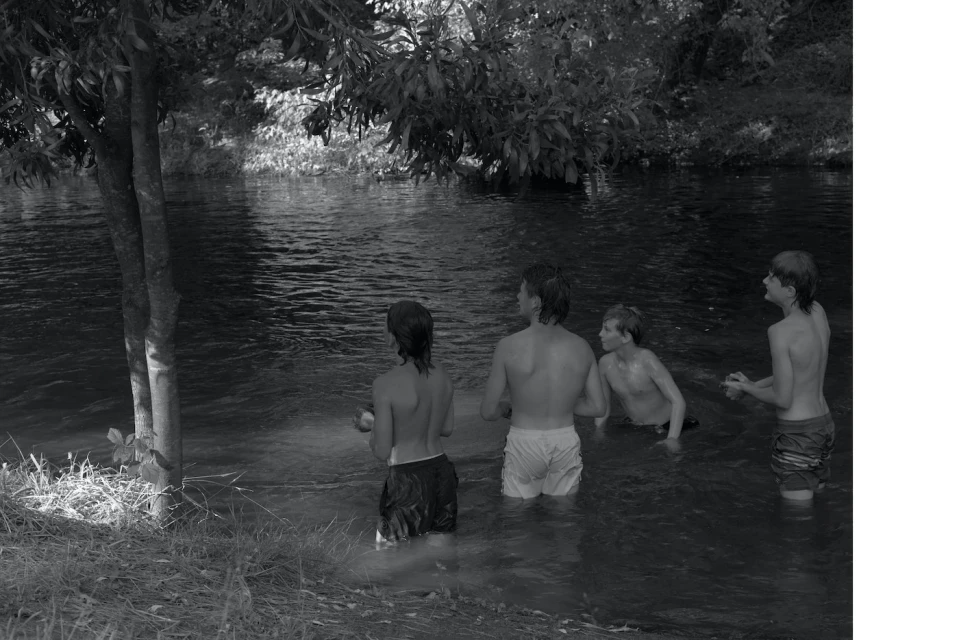 Four youths stand in a river surrounded by trees and vegetation, captured in a monochromatic image with a documentary feel.