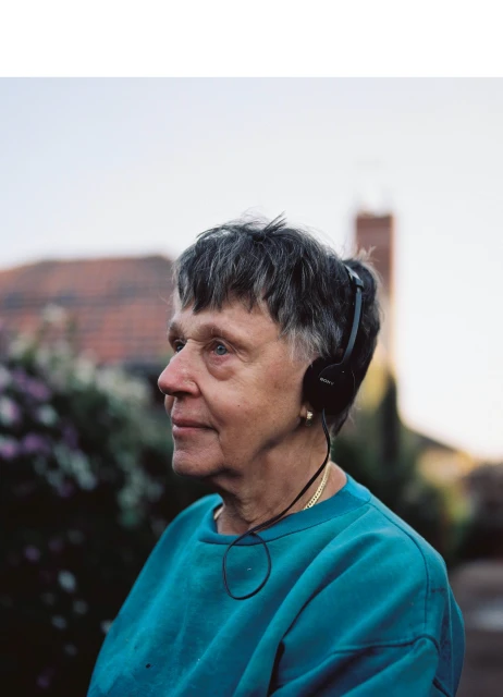 Elderly woman wearing headphones and a blue sweater stands outside in front of a brick building and green foliage with a light blue sky.