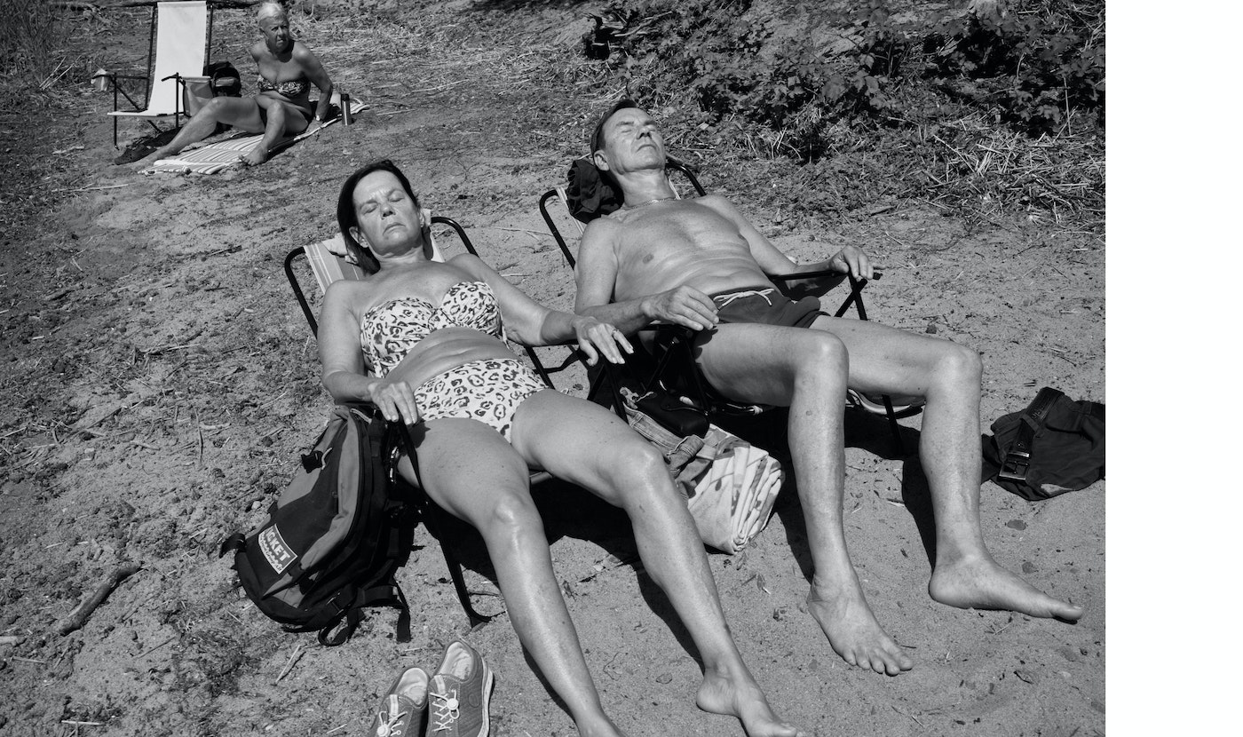 Black and white shot capturing two adults relaxing on beach chairs under sunlight near foliage with another sunbather in the background.