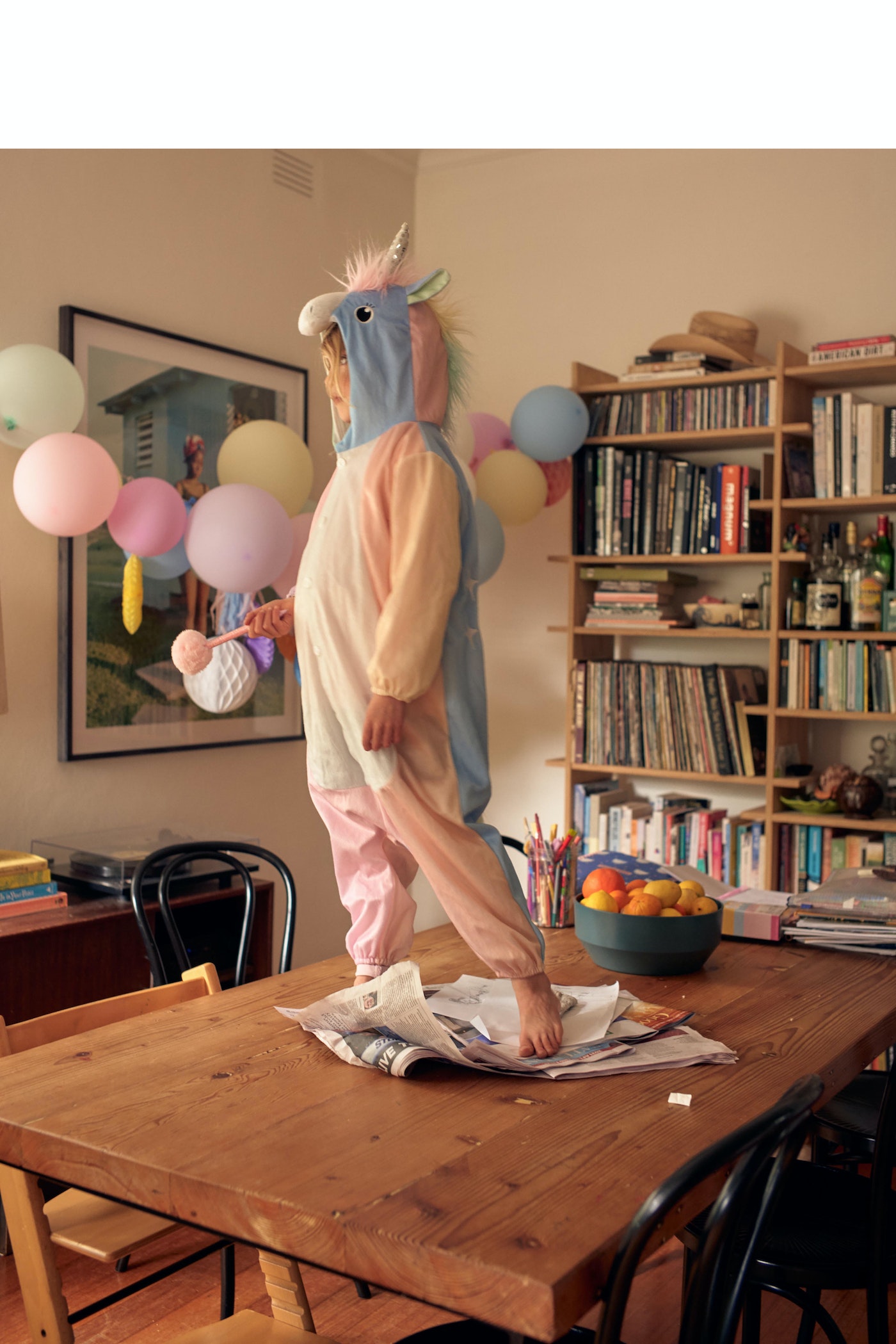 Child in unicorn costume stands on a wooden table surrounded by balloons and books in a brightly lit indoor setting with a playful mood.