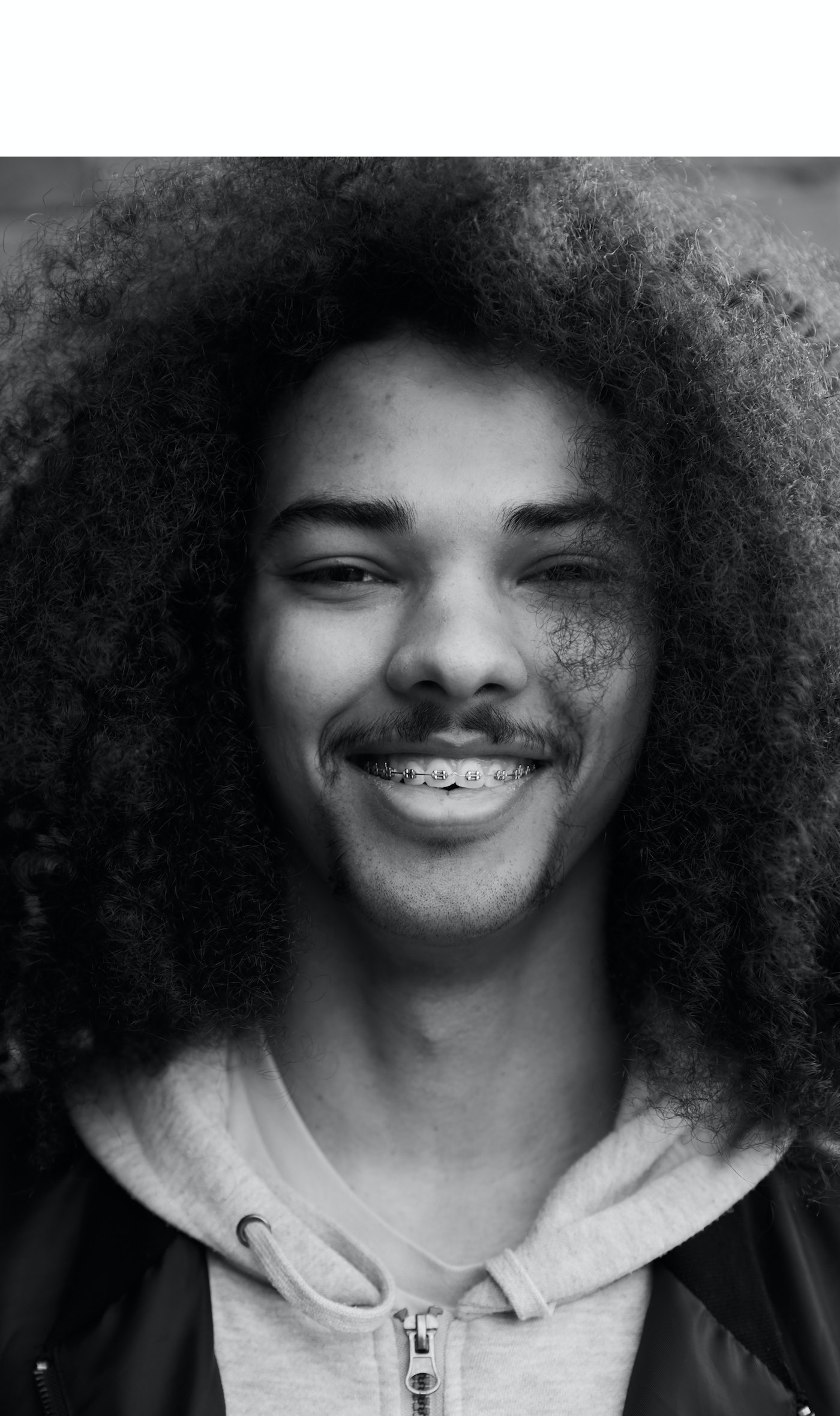 Close-up portrait of a young adult male with an afro and braces, wearing a zip-up hoodie, set against a neutral blurred background.
