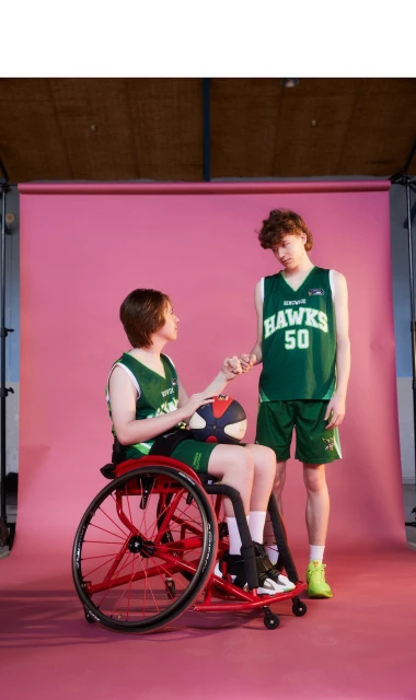 Two young men in basketball uniforms, one in a wheelchair, pose in a studio against a pink background, holding hands and looking at each other.
