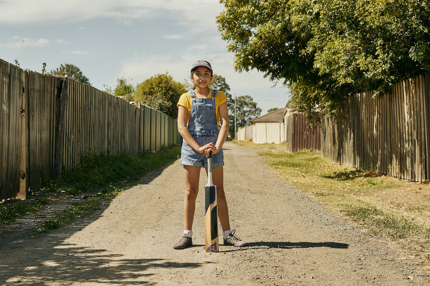 Young girl stands confidently with cricket bat on a dirt path lined with fences, trees, and buildings under a bright sunny sky.