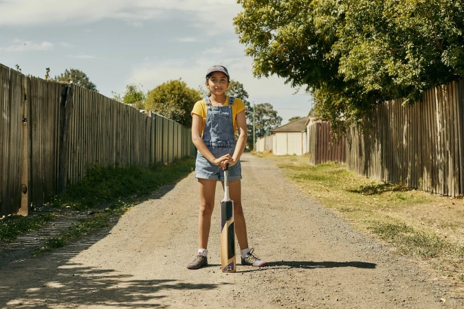 Young girl stands confidently with cricket bat on a dirt path lined with fences, trees, and buildings under a bright sunny sky.