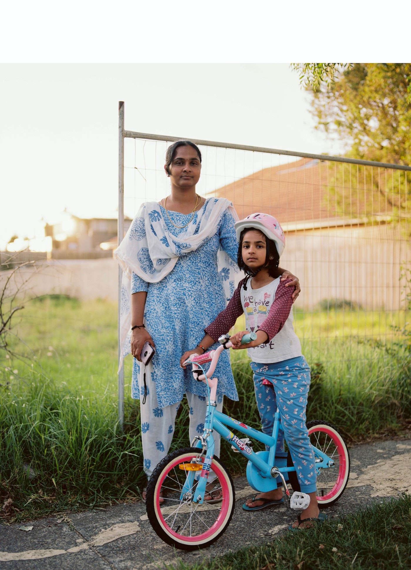 A mother and daughter stand next to a bicycle on a sunny day; the young girl is wearing a helmet and posing with the blue bike.