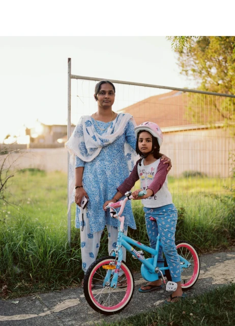 A mother and daughter stand next to a bicycle on a sunny day; the young girl is wearing a helmet and posing with the blue bike.