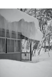 Monochrome photograph of a cabin roof heavily covered in snow with long icicles hanging over a porch.