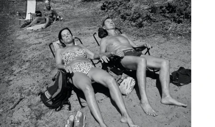 Three people relaxing in folding beach chairs on sandy ground under bright daylight, captured in monochrome.