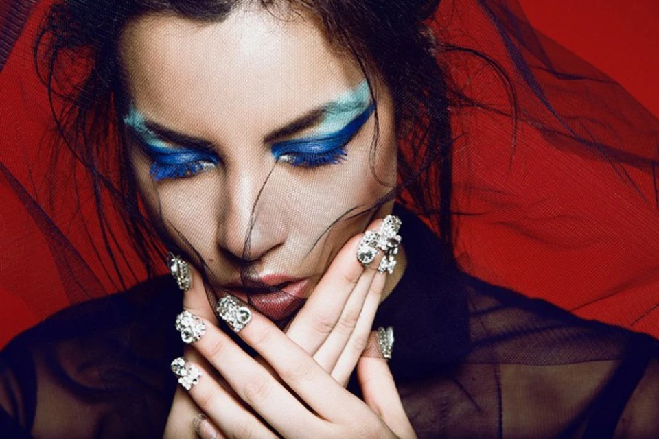 Close-up of a woman with dramatic blue eye makeup, wearing a black veil and multiple ornate silver rings, against a red backdrop.