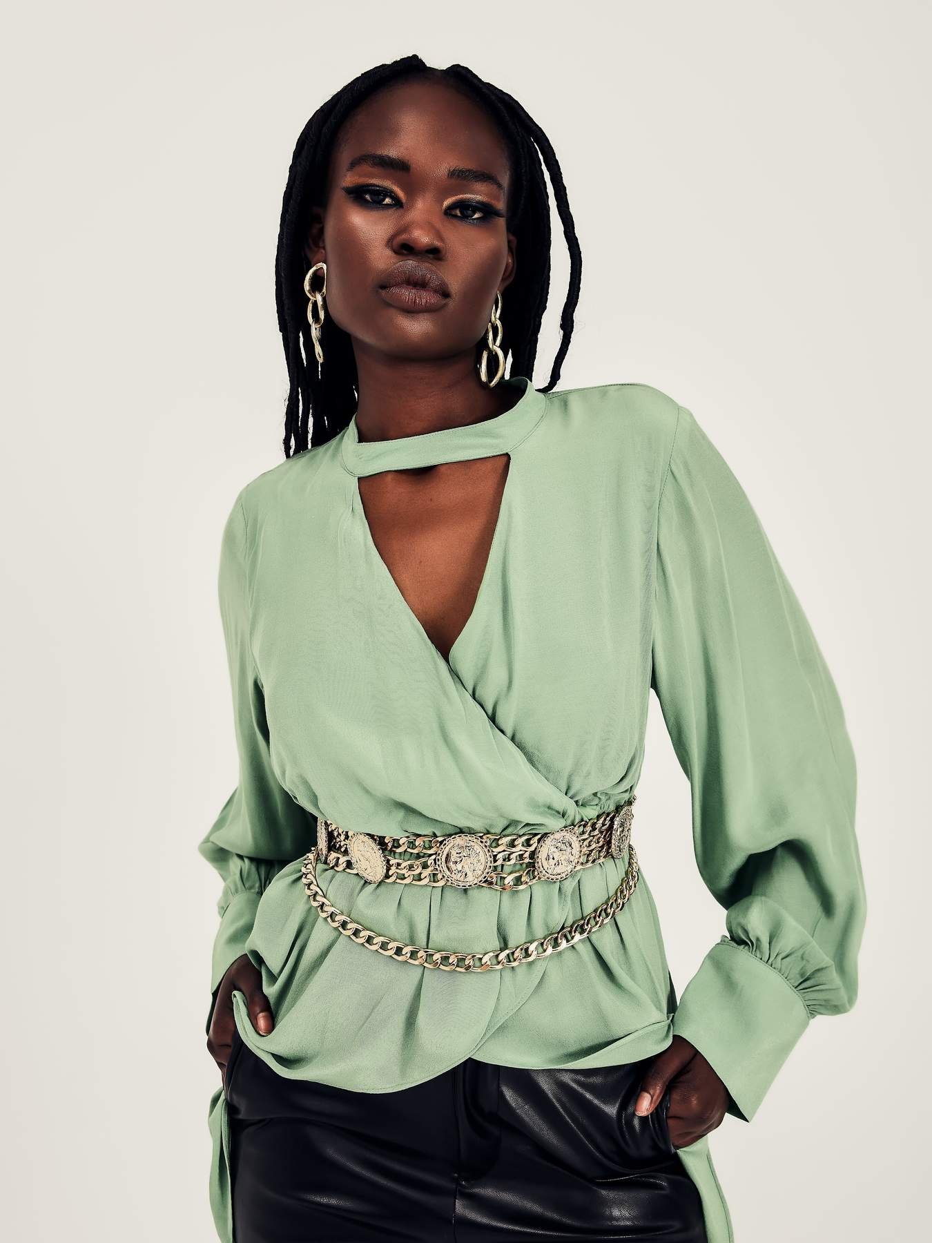 An African-American woman is in a light-green blouse and coin-studded belt against a white backdrop, with long dark hair, jewelry, and dark eyes.