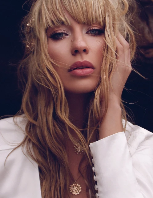 Close-up of a blonde woman wearing a white jacket and gold necklaces, gazing at the camera with a neutral expression in an indoor setting.